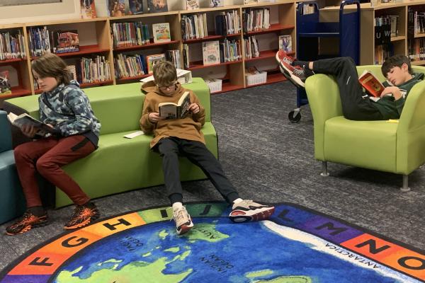 three boys reading books in a school library while sitting on furniture