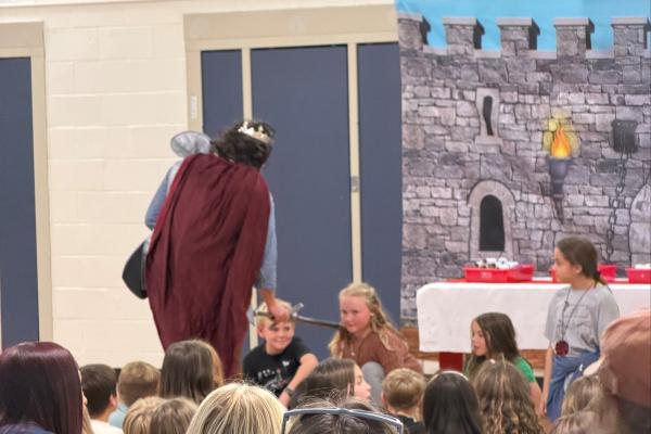a king (school principal) and a 4th grade girl with long blonde hair  and other classmates being "knighted" during a middle ages day knighting ceremony. 