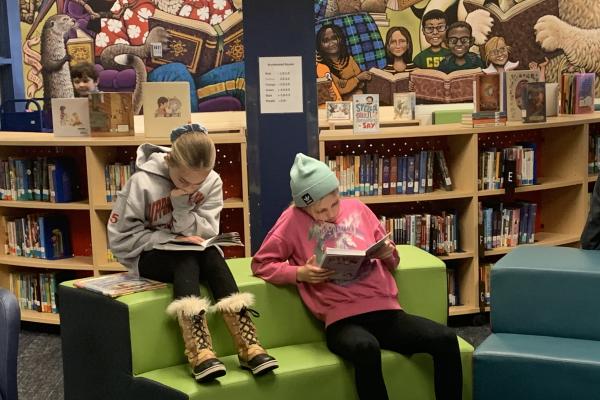 two girls reading books while sitting on furniture in the school library