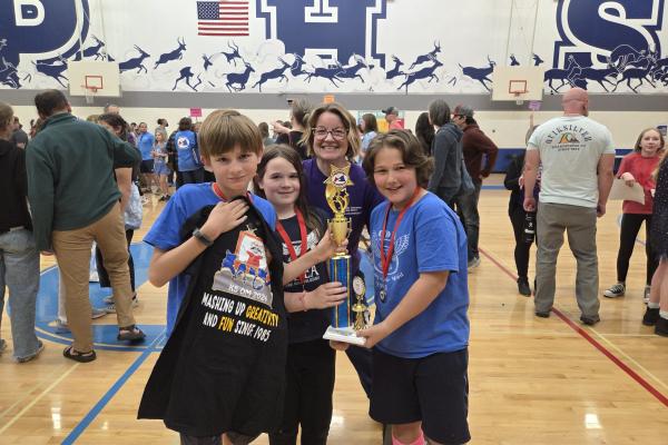 two boys and a female teacher smiling and holding a trophy after winning an OM tournament