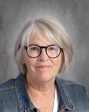 headshot - smiling female teacher with gray hair and glasses wearing a white top with a blue jacket