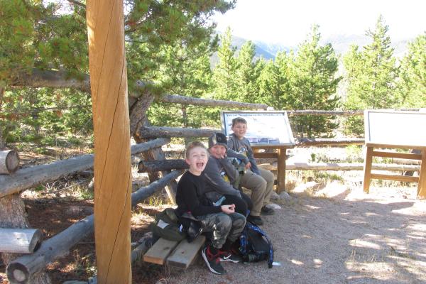 three boys at Colorado State Universite Mountain Campus on ECO week trip sitting at a wooden table surrounded by pine trees smiling 