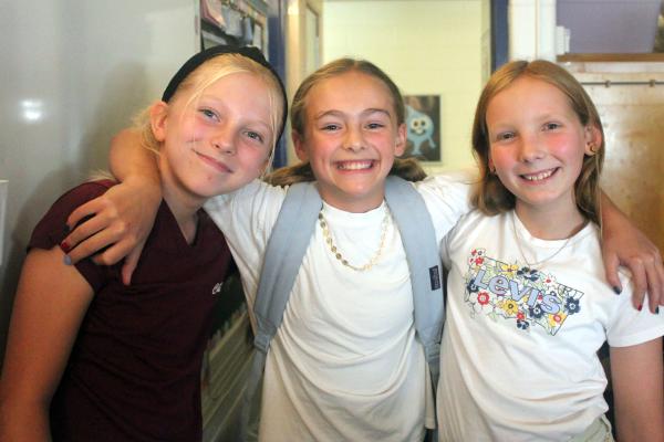 Three girls with arms around each other's shoulders smiling, happy to see each other at meet your teacher night.