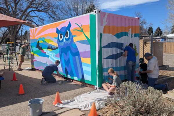 five parents - men and women - working on painting a mural and attaching an owl with sunset and mountains  mural to a shipping container