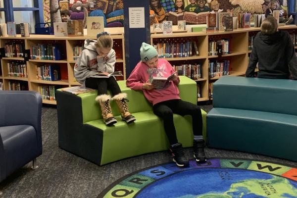 two girls reading books while sitting on furniture in the school library