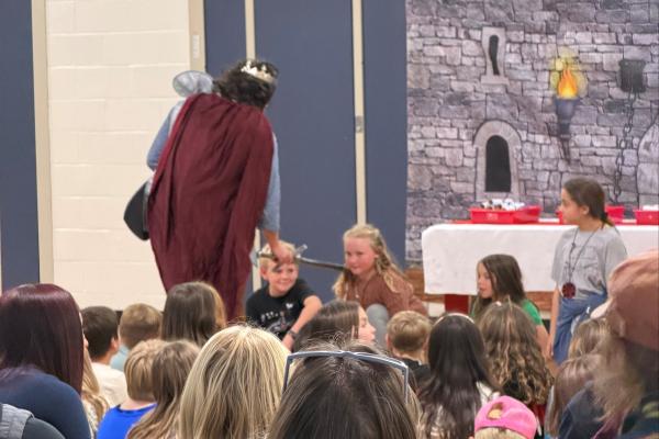 a king (school principal) and a 4th grade girl with long blonde hair  and other classmates being "knighted" during a middle ages day knighting ceremony. 